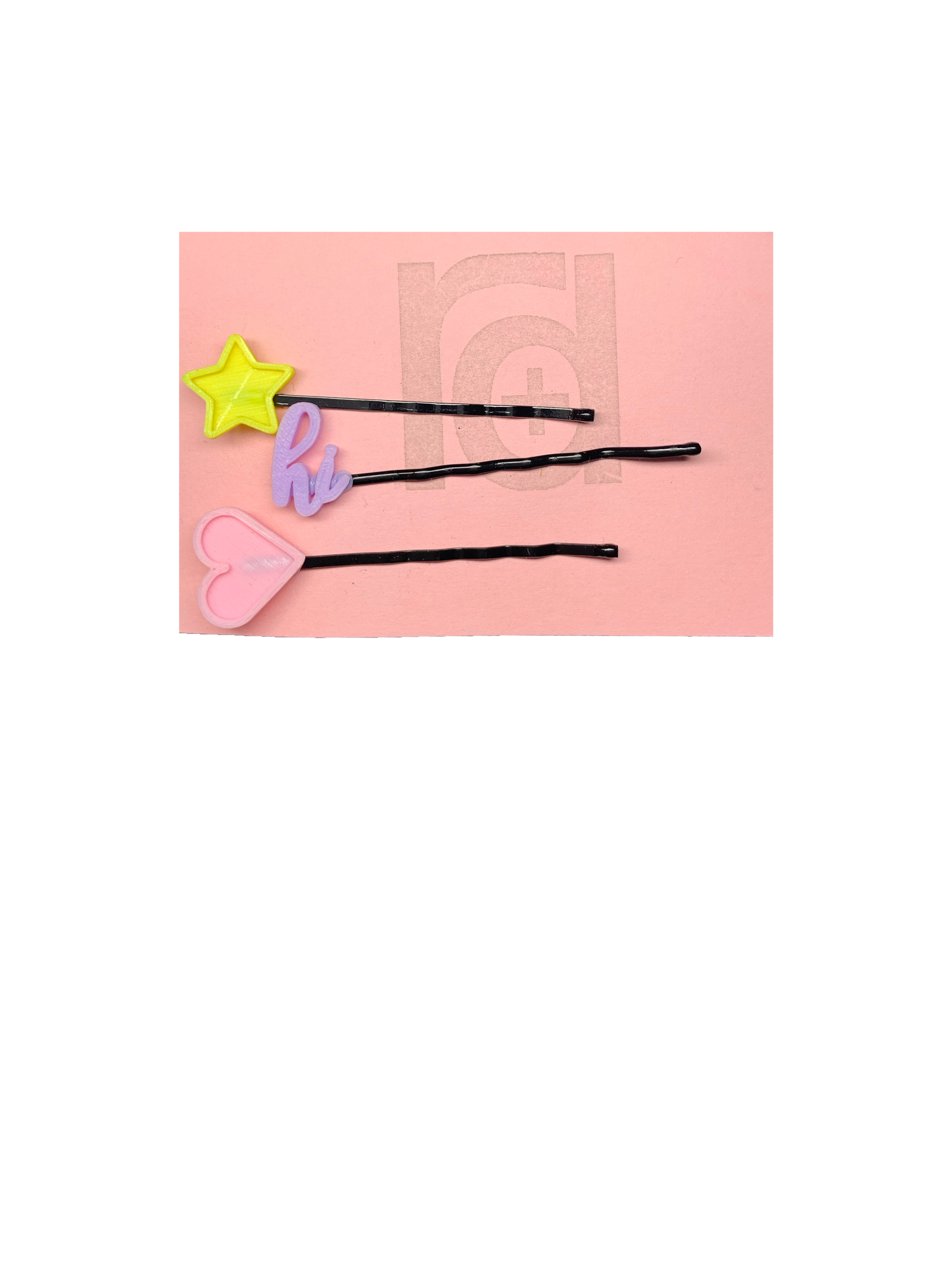 On a pink card are three bobby pins with 3D printed charms on the end from R+D. The  top bobby pin has a bright yellow star. The second is light purple and in cursive script says hi. The third is a light pink heart. All of the packaging is eco friendly, either recyclable or compostable.