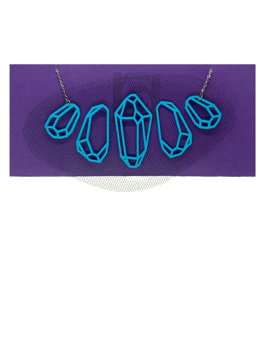 On a bright purple necklace paper is a statement necklace from R+D. It features 5 gems in a symmetrical formation that get larger in the center.  The 3D printed gems are embedded with tulle fabric. They give the effect of a floating necklace. This particular necklace has black tulle embedded in teal filament. 