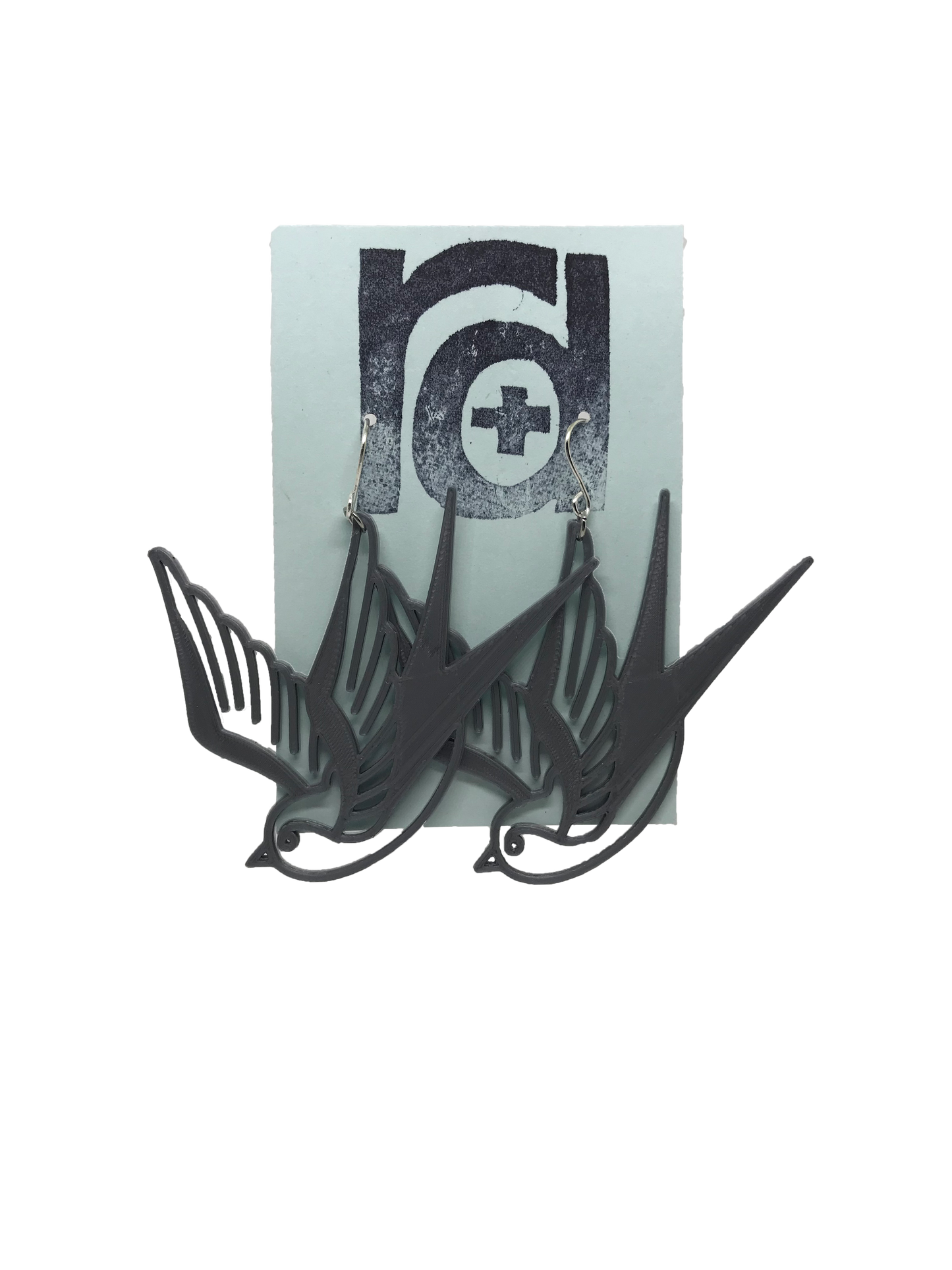 On a light blue earring card are two R+D earrings. They are an eco friendly silver and look like two swallow birds that are circling or flying down.