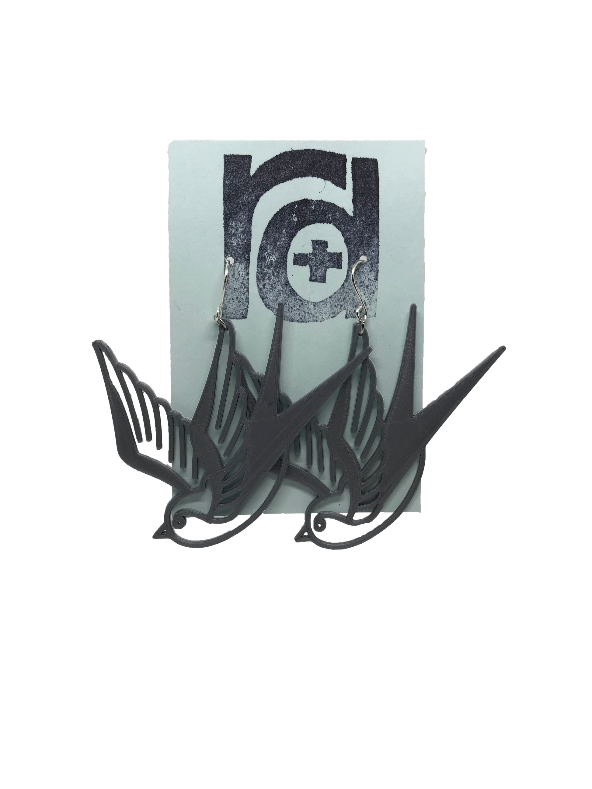 On a light blue earring card are two R+D earrings. They are an eco friendly silver and look like two swallow birds that are circling or flying down.