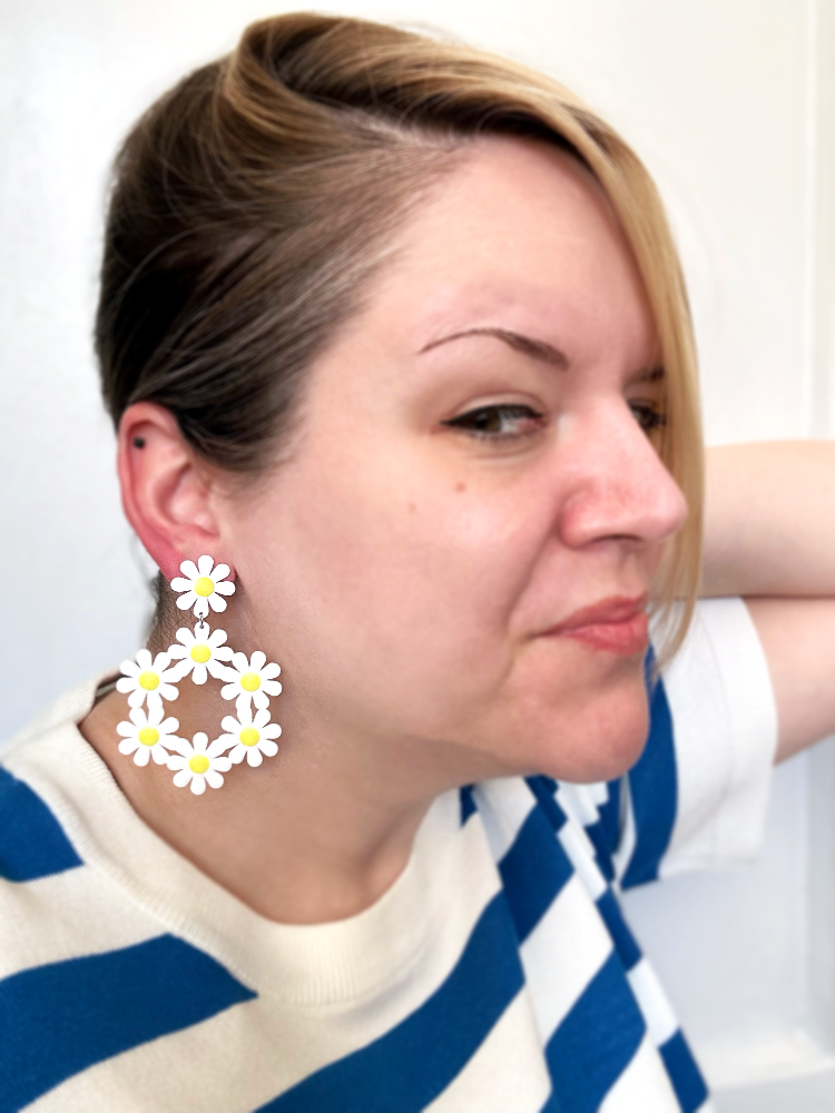 This photo shows someone pulling their hair to one side to show an earring hanging from their lobe. They are wearing a 3D printed earring that is a single white petal daisy with a yellow center as the stud. Below 6 more white and yellow daisies form a circle wreath.