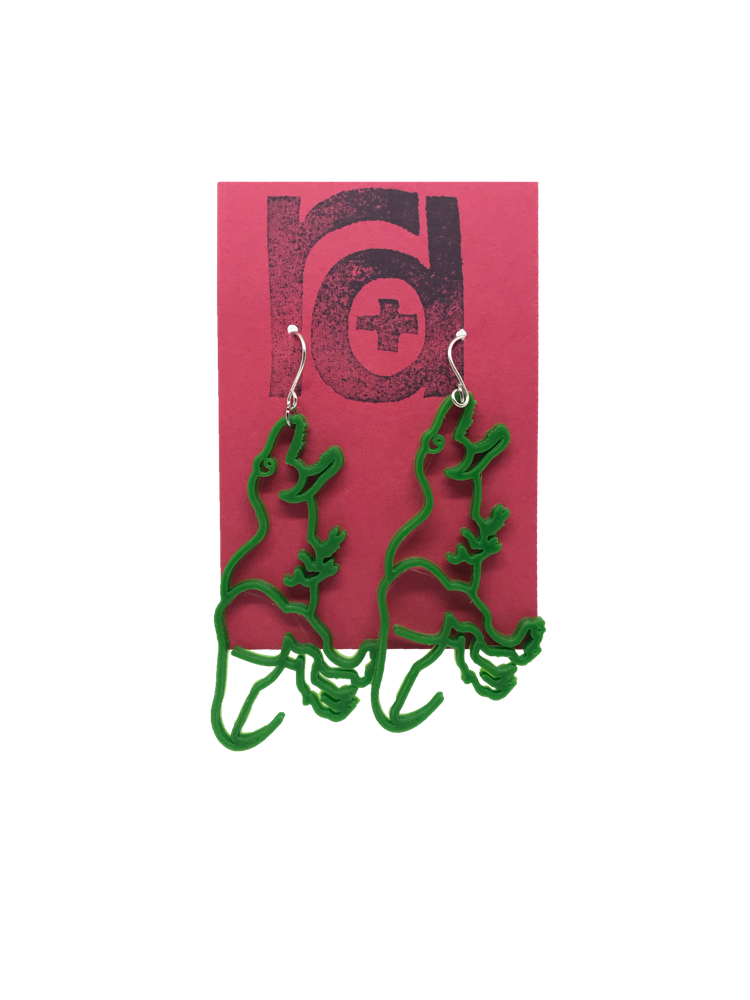 On a red R+D earring card is a pair 3D printed earrings. They are shaped as dinosaurs, Tyranasaurus Rex specifically. They are a bright kelly green color and hang from the hook in a way that looks as though the t-rex's open mouth is headed to its the wearer's earlobe.