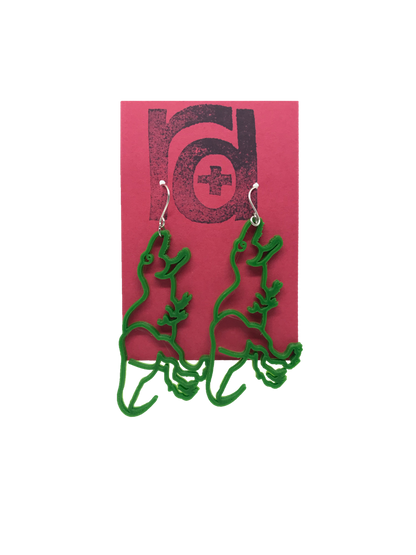 On a red R+D earring card is a pair 3D printed earrings. They are shaped as dinosaurs, Tyranasaurus Rex specifically. They are a bright kelly green color and hang from the hook in a way that looks as though the t-rex's open mouth is headed to its the wearer's earlobe.