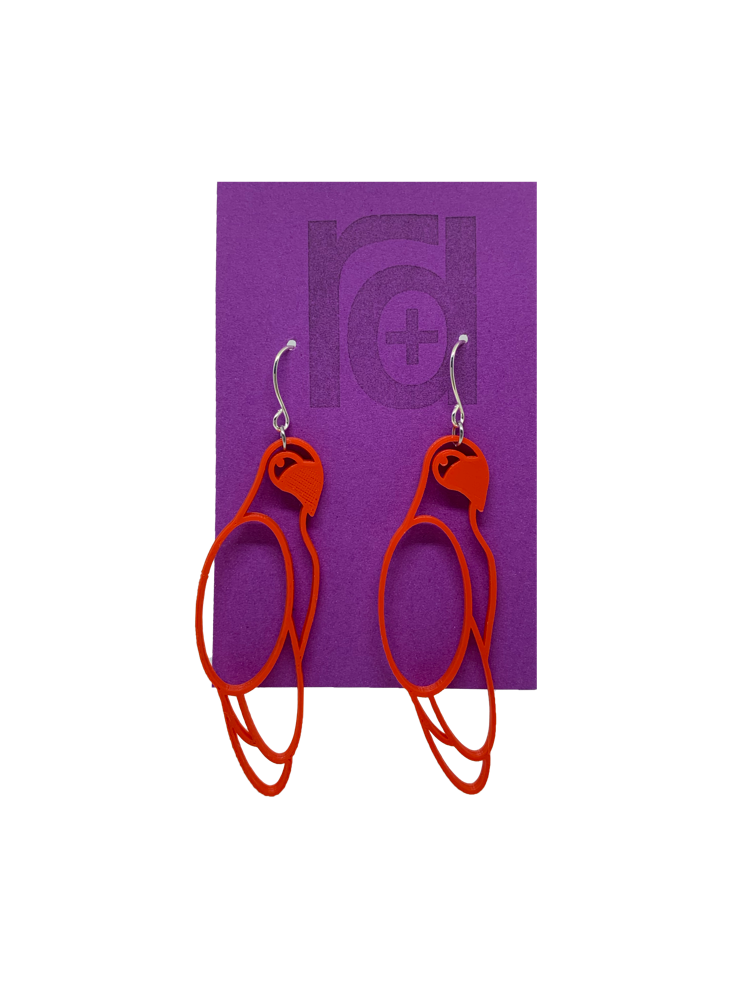 Hanging from a bright purple earring card are two R+D 3D printed earrings. The earrings are red outlines of parrots.