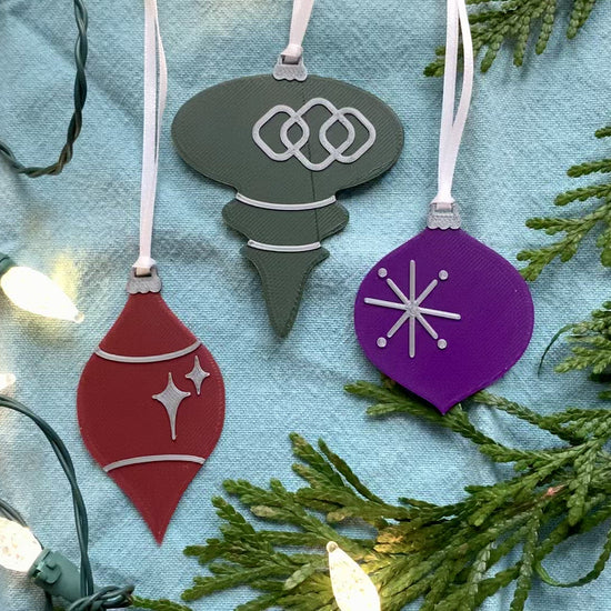 On a bright blue background with  evergreen branches and a string of lights are a set of 3 R+D 3D printed ornaments. They are all designed to look like vintage baubles that would have commonly been found on Christmas trees in the past. They each have silver accents of stars and shapes. One is merlot red, one is dark green, and one is purple.