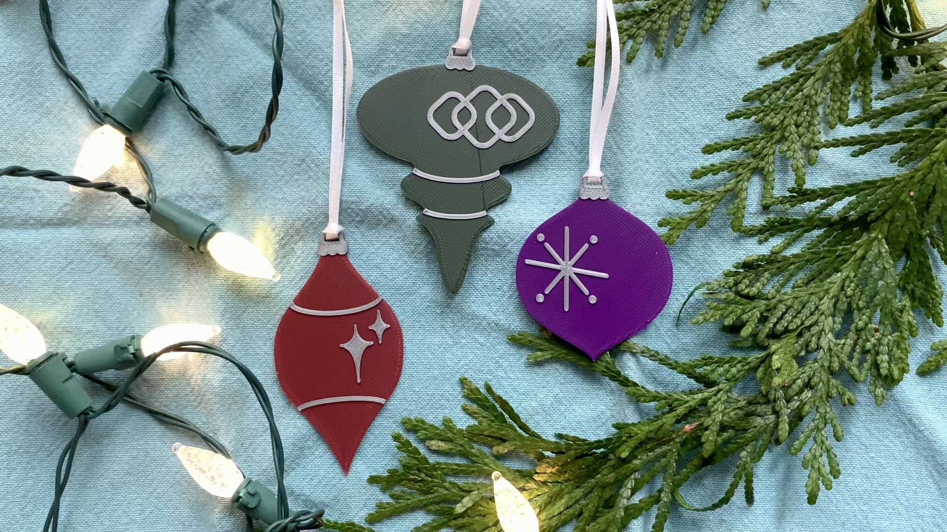 On a bright blue background with  evergreen branches and a string of lights are a set of 3 R+D 3D printed ornaments. They are all designed to look like vintage baubles that would have commonly been found on Christmas trees in the past. They each have silver accents of stars and shapes. One is merlot red, one is dark green, and one is purple.