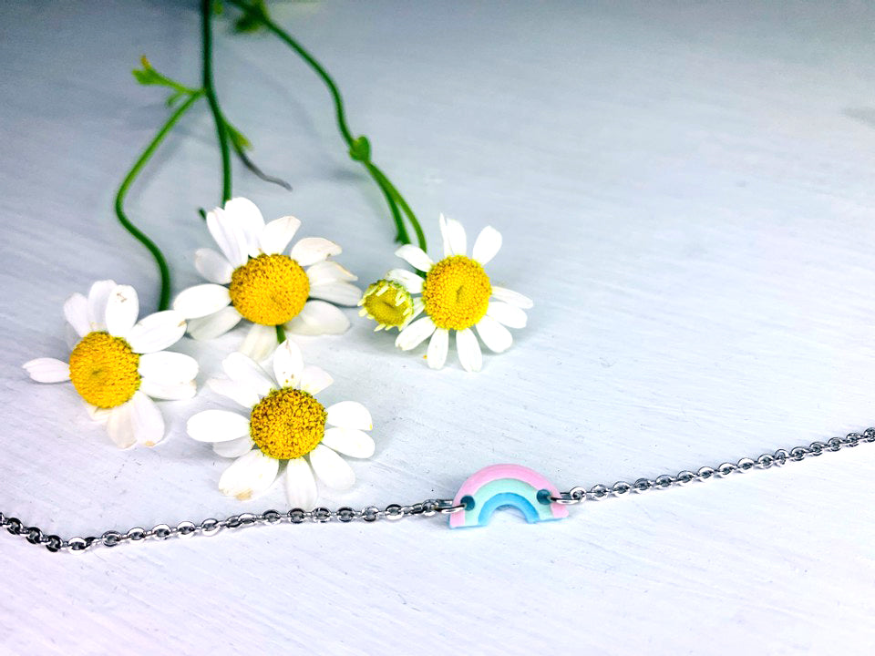On a white background is a bracelet laid out in front of white and yellow chamomile flowers. The bracelet features a charm in the middle that is a small 3D printed rainbow made from pastel pink, mint green, and baby blue. 