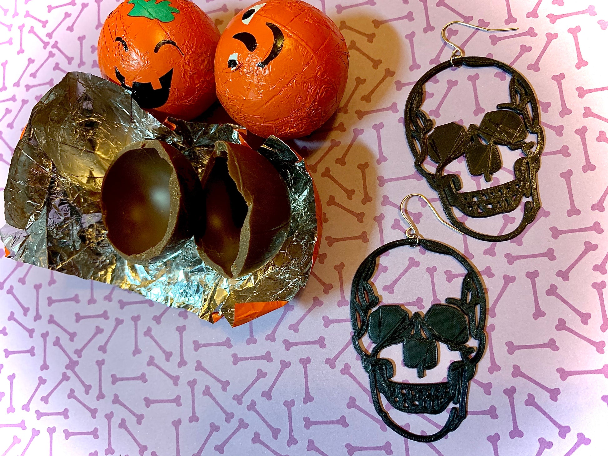 Two earrings are shown with pumpkin chocolates for halloween. The earrings are black and in the shape of two large human skulls.