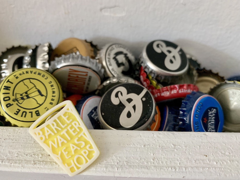 In front of a pile of beer tops is a 3D printed pin. It looks like a pint glass and has the words barley, water, yeast, and hops incorporated in, which are the four main ingredients of beer. 