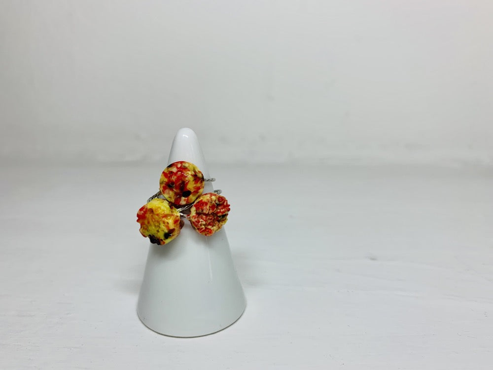 On a white background and slipped on a white cone ring holder are three rings cast from recycled 3D prints. The rings are each in a circular shape. One has a smooth surface, another a faceted surface, and finally one like a geode forming. They each have red, yellow, orange, and black colors intermixed to be speckled like granite.
