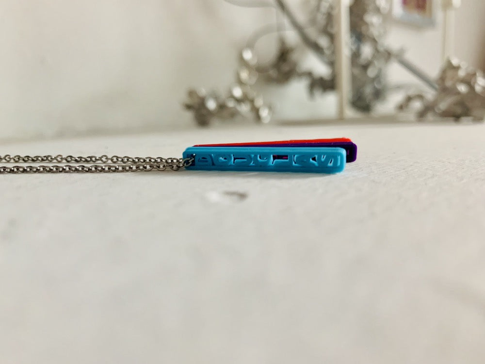 On a white background are three 3D printed pendants. They are each different colors and shaped as long rectangles with names in the center. When worn, they simply look like long hanging rectangles, but from the side, all the names are visible. This one has a teal pendant, a purple pendant, and a red pendant, but from this perspective the  teal pendant is most visible. It reads Douglas. 