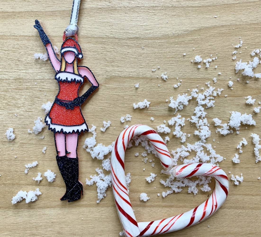 On a wood background with white snow and candy canes shaped into a heart, there is a 3D printed ornament from R+D. The ornament is printed in a plant based filament. It is shaped like Regina George from the movie Mean Girls. She is striking the iconic pose at the beginning of performing Jingle Bell Rock, wearing black gloves and boots and a red outfit with white trim. The entire ornament is covered in glitter to shimmer and shine in the light.