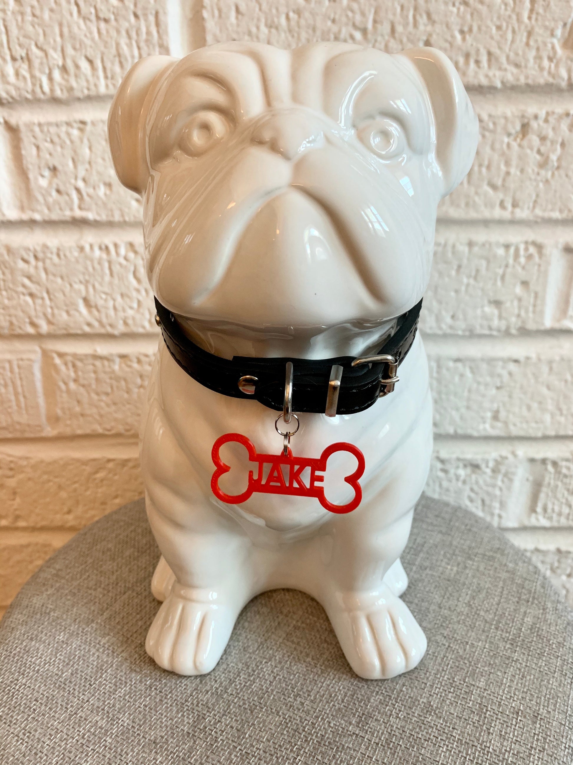 In front of a white brick wall and on a grey cushion is a ceramic bulldog statue. It is wearing a black collar and has a red dog tag. The dog tag is 3D printed in the shape of a dog bone with the name JAKE in the middle. The name can be personalized to any name you would like. 
