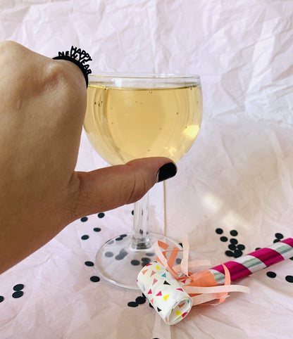 Reaching into the frame is a hand with a 3D printed ring on the pointer finger. It is a very small and delicate ring that reads Happy New Year. It looks like the paper crowns people wear on New Years Eve. The hand is reaching for a glass of champagne and a colorful noisemaker and black confetti are resting nearby. 
