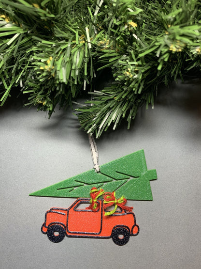On a grey background and hanging from a green wreath is a R+D 3D printed ornament hanging down. It is shaped as vintage style red car with a big green tree strapped on top. It is held together with red and green bows. The entire ornament is covered in glitter to be able to shimmer and shine in the light.