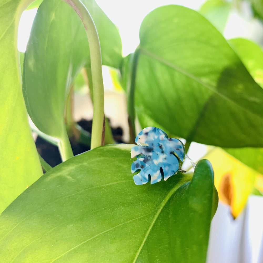 On bright green leaves is a ring with a thin twisted band and a charm. The charm is the shape of a monstera leaf with teal, blue, white, and black colors intermixed to be speckled like granite.