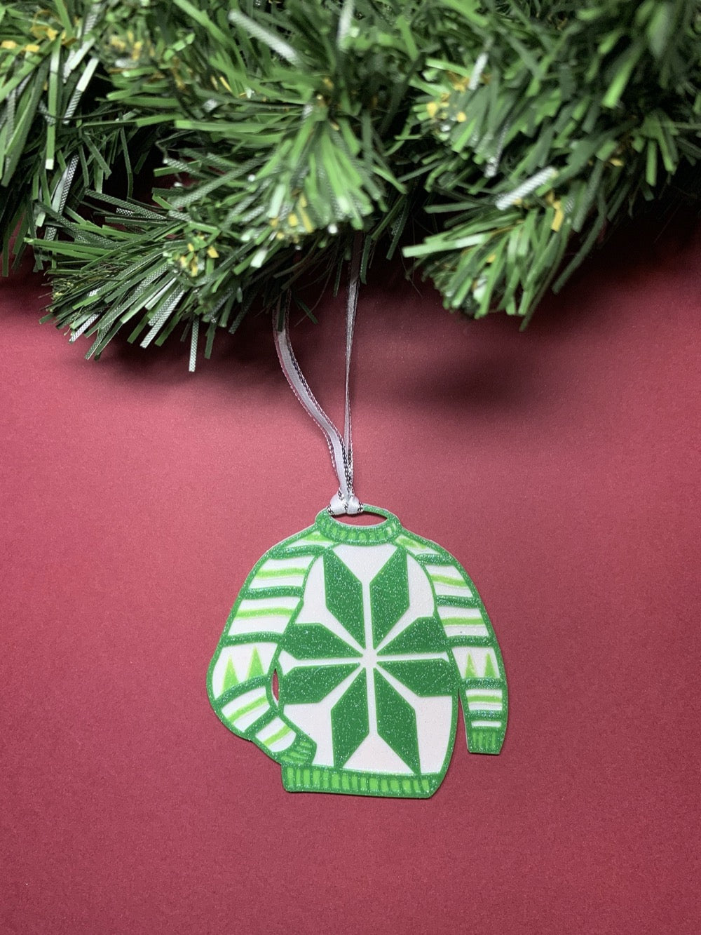 Shown on a red background and hanging from a wreath is a R+D 3D printed ornament. It is in the shape of an ugly sweater with green and white stripes, trees, and a snowflake.