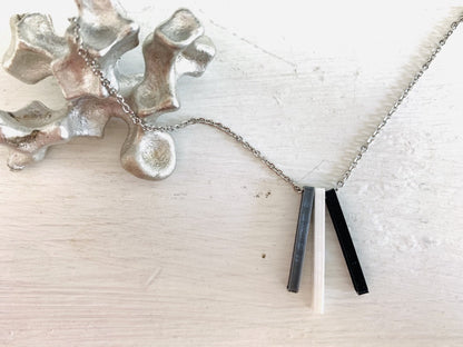 Shown on a white background with an aluminum sculpture on the side. There is a necklace with 3 3D printed pendants. The pendants are long and thin, in silver, white, and black. When turned to the side, names are visible on the pendants.