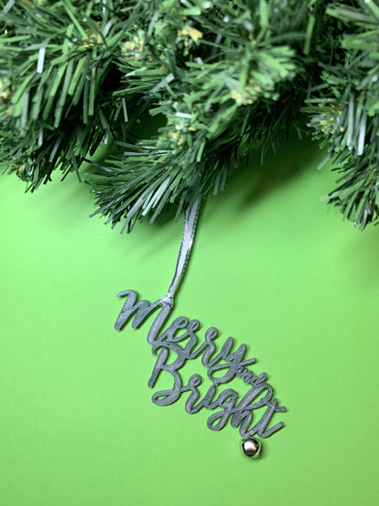 On a bright green background and hanging from a green wreath is a 3D printed R+D ornament. It is a cursive text with a jingle bell and covered in glitter to make it shimmer and shine in the light. This ornament reads, Merry and Bright.