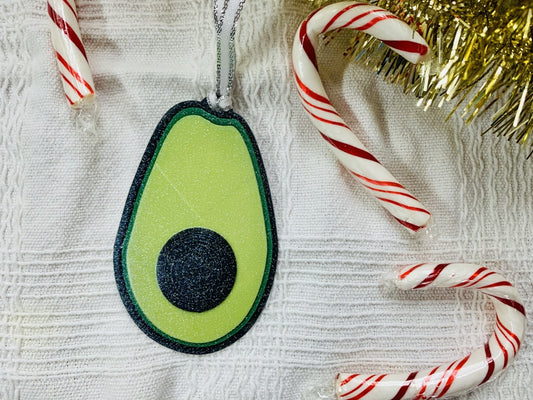 On a white fabric background is a R+D 3D Printed ornament. It is shaped like an avocado that has been split open with a black peel, bright green highlights and lime green in the center. There is also a raised black pit in the middle. The entire ornament is covered in glitter so it will shine and shimmer in the light. To the side there are also red and white candy canes and gold garland. 