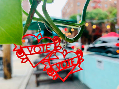 In the foreground, hanging off of a green vine are two bright red 3D printed earrings. The earrings are heart shapes with a banner twisted around them and a rose on each side. Across the banner it reads vote, which can be customized with any word. 
