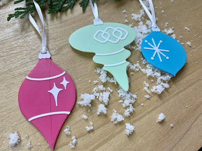 On a wood background with white snow and hanging from an every green branch are a set of 3 R+D 3D printed ornaments. They are all designed to look like vintage baubles that would have commonly been found on Christmas trees in the past. They each have white accents of stars and shapes. One is hot pink, one is mint green, and one is teal.