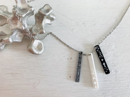 Shown on a white background with an aluminum sculpture on the side. There is a necklace with 3 3D printed pendants. The pendants are long and thin, in silver, white, and black. These are turned to the side to reveal three names: SEAMUS, LUCIUS, and REBEKAH. They can be customized to any name or word. 