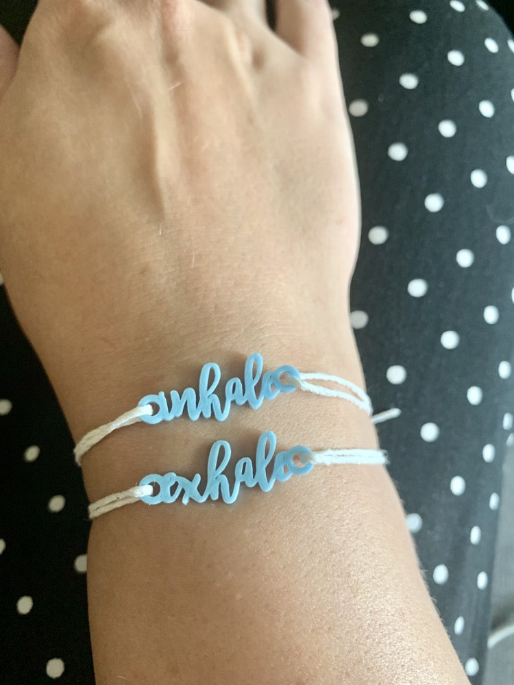 Shown on a wrist are two 3D printed charm bracelets. They are light blue with white cotton adjustable cords. They are both in cursive. One says inhale, one says exhale.