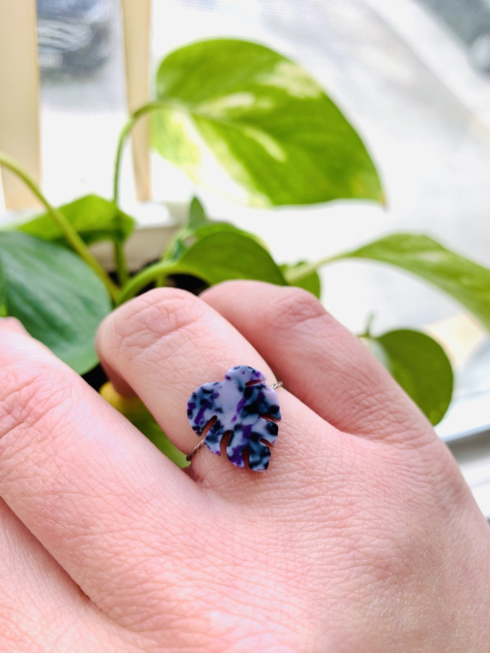 In the foreground is a hand wearing a cast ring in the shape of a monstera leaf. It is cast from recycled 3D prints in hues of purple, black and white. In the background are bright green leafs. 
