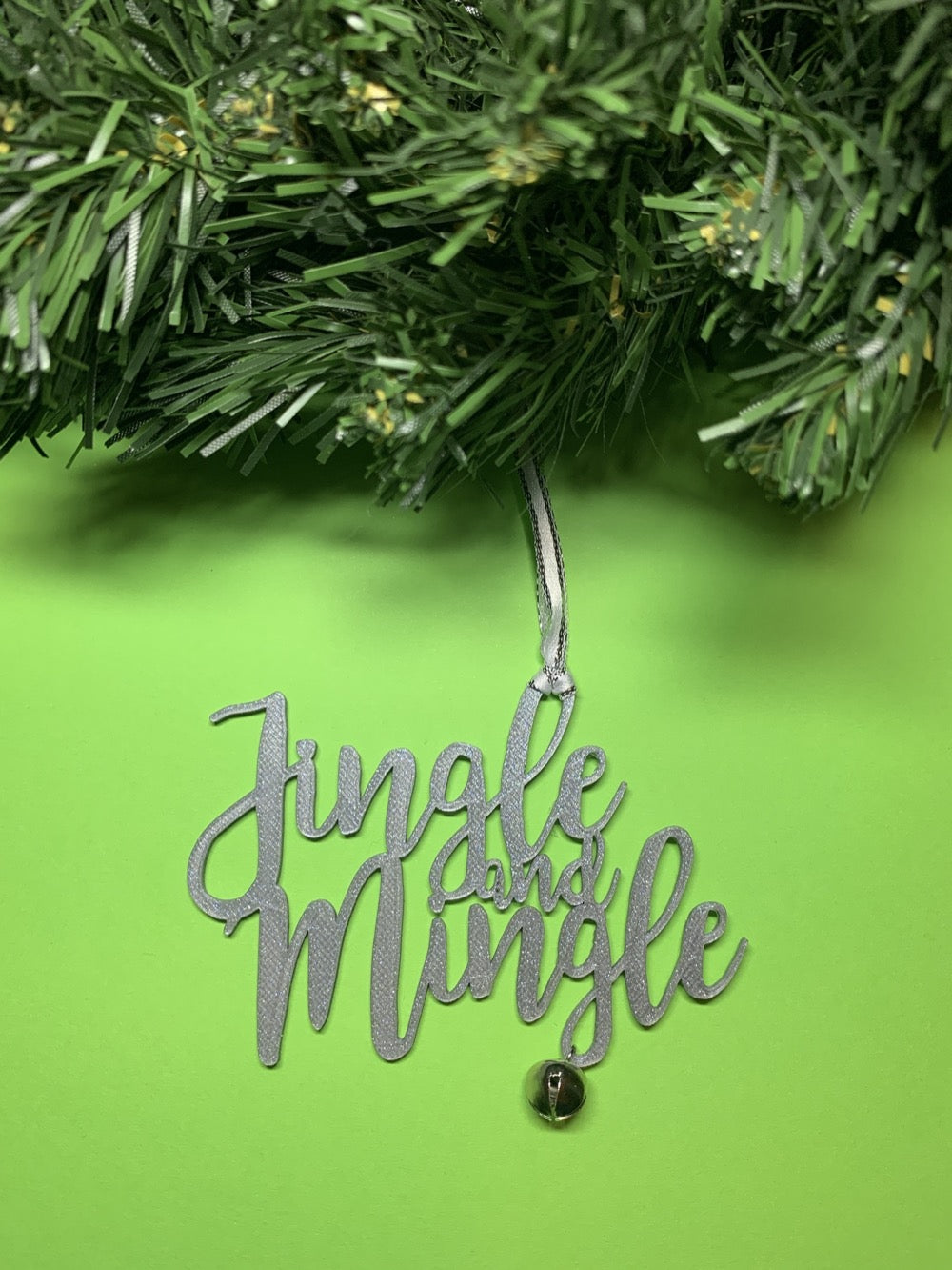 On a bright green background and hanging from a green wreath is a 3D printed R+D ornament. It is a cursive text with a jingle bell and covered in glitter to make it shimmer and shine in the light. This ornament reads, Jingle and Mingle. 