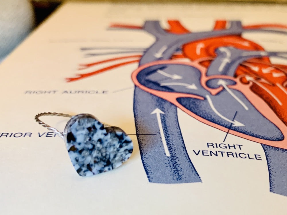 Laying on an old textbook with a drawing of a human heart is a ring that is cast from recycled 3D prints and scraps. The gem on the front is a heart with a smooth surface. It incorporated black, white and silver colors creating a speckled look like granite. 