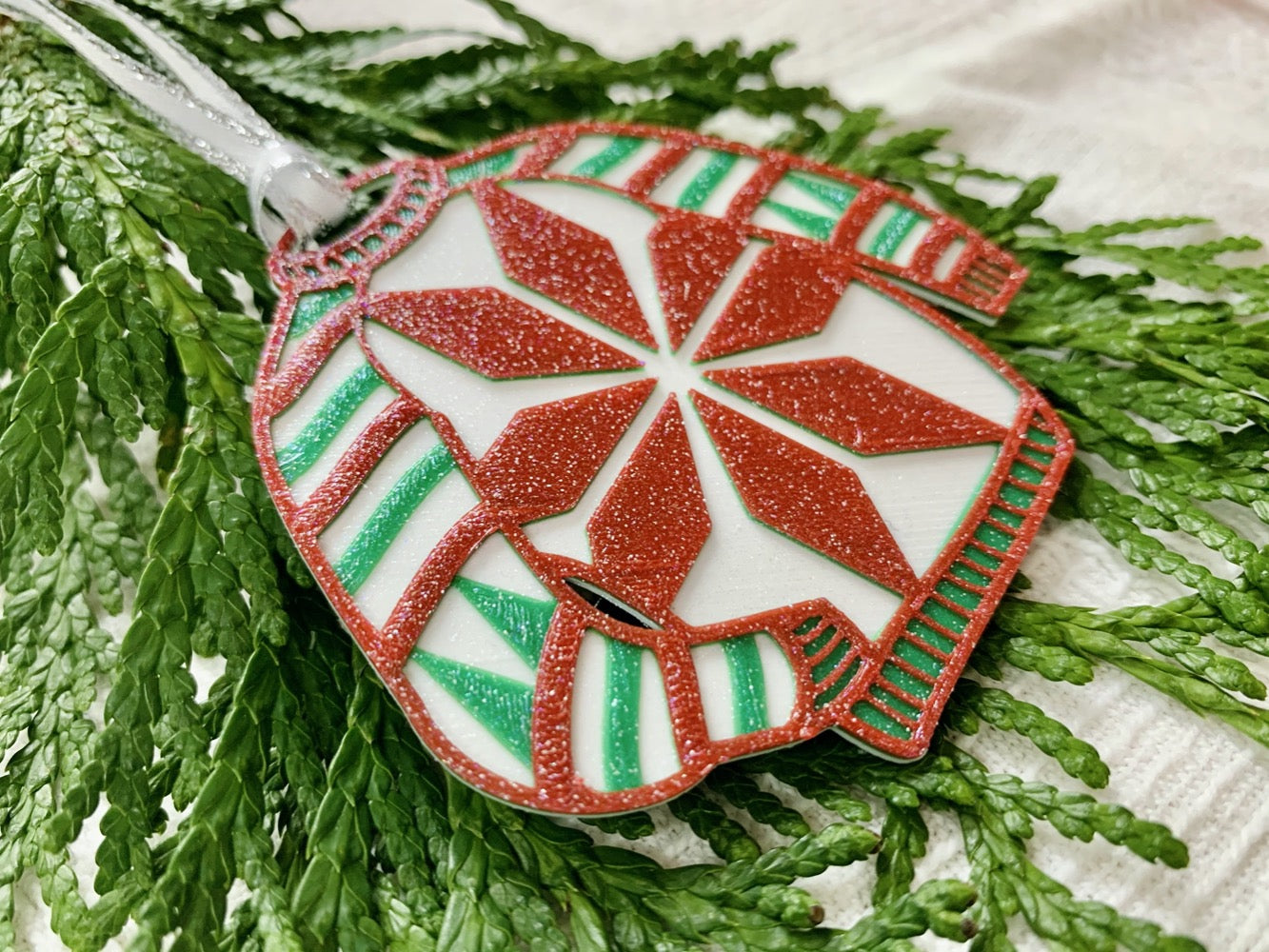 Shown on a white fabric background and with sprigs of evergreen is a R+D 3D printed ornament. It is in the shape of a chunky holiday sweater. It is white with red and green stripes, trees on the sleeves, and a snowflake in the center. The entire ugly sweater ornament is covered in glitter so it will shimmer and shine in the light.