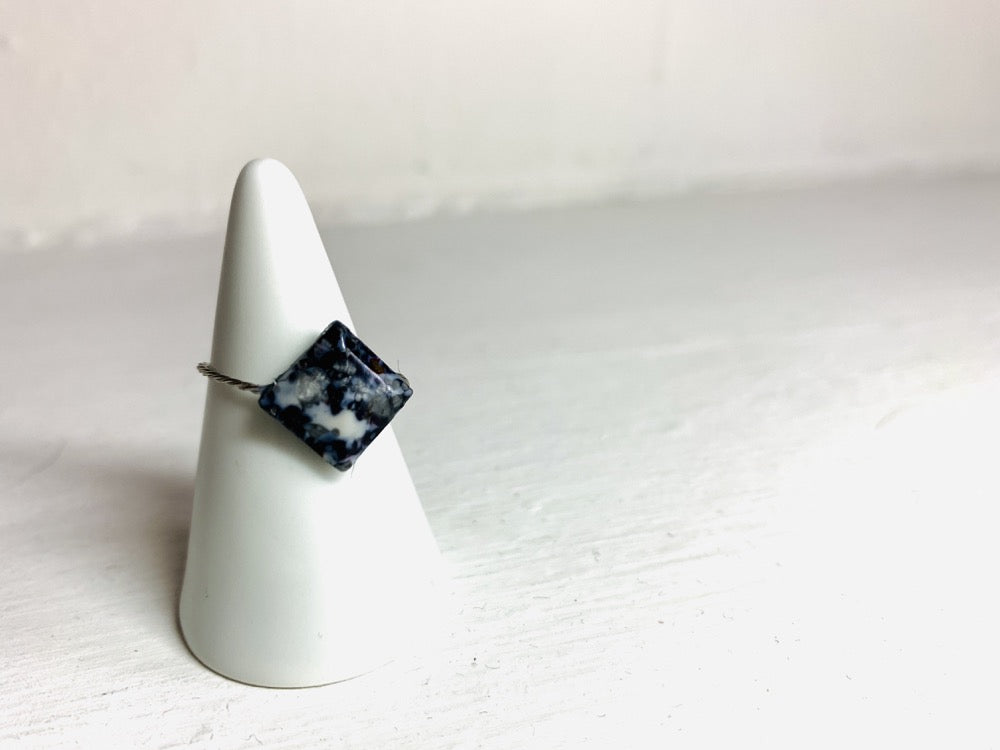 On a white background and slipped on a white cone ring holder is a ring that is cast from recycled 3D prints. The ring is in a faceted square shape with black, white, and silver colors intermixed to be speckled like granite. 