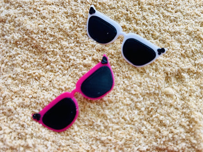 Shown in sand are two 3D printed pins from R+D. They are both shaped like cat eye sunglasses with black lens and black circle accents in the upper corners. The frames are two different colors. One is a bright pure white and the other is a hot pink.
