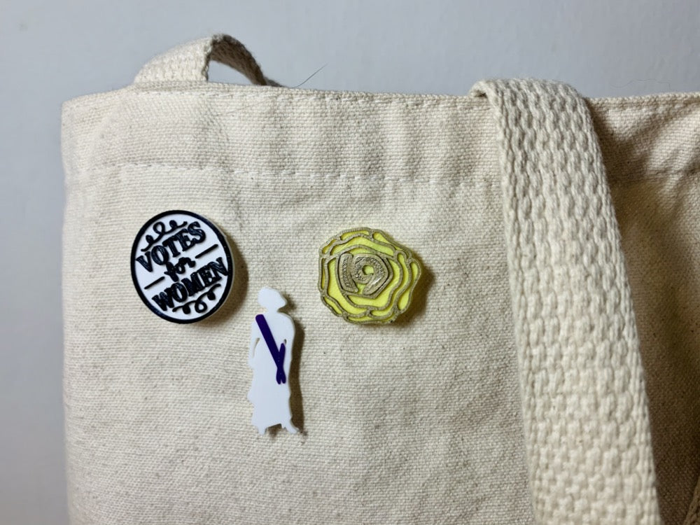 Shown on a tan tote bag are three R+D 3D printed pins. The first on the left is a white circle with a black outline and words saying "Votes for Women". The next is a silhouette of a woman from the 1920s. Aside from the silhouette, there is a purple sash. The third pin is a yellow rose, which was worn in support of the movement. The petals are all outlined in gold and in the center is the number 19 for the 19th amendment.