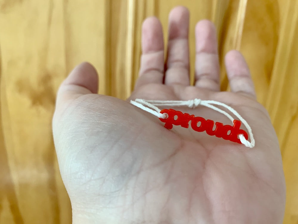 Shown on an outstretched hand is a bracelet with a 3D printed charm. The adjustable cord on the bracelet is a white organic cotton. The charm is bright red and says proud in block letters. 