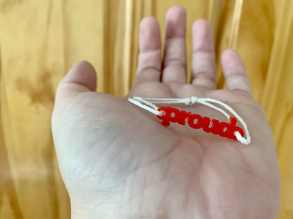 Shown on an outstretched hand is a bracelet with a 3D printed charm. The adjustable cord on the bracelet is a white organic cotton. The charm is bright red and says proud in block letters. 