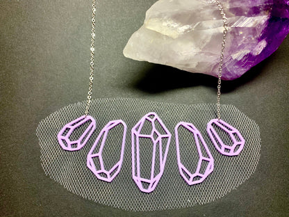 On a black background is a R+D 3D Printed statement necklace. The five light purple gems symmetrical on the necklace and are embedded with white tulle and a chain is resting over a purple and white amethyst crystal.