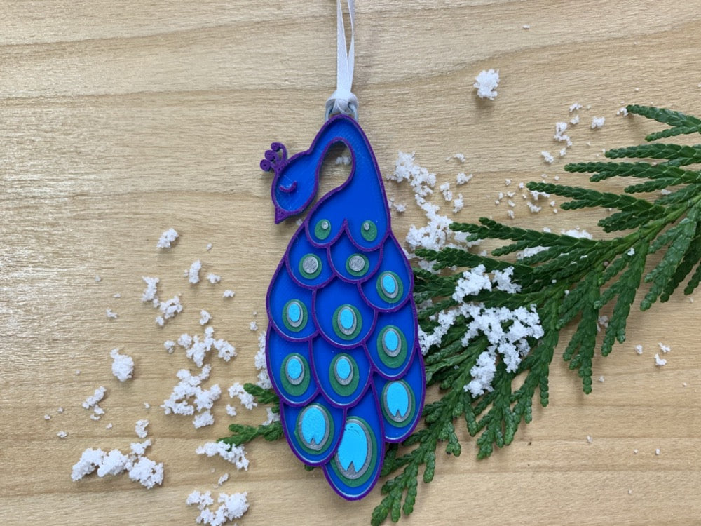 On a wooden shelf is an evergreen sprig, white snow, and a R+D 3D printed peacock ornament. It has a blue body, with purple outlines. On the long elegant feathers are green, silver and teal accents.