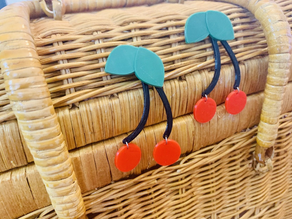 On the side of a wicker picnic basket are two R+D 3D printed earrings. Each earring has a two bright red cherries at the end of two connected black stems. The top of the earring has kelly green leaves. 