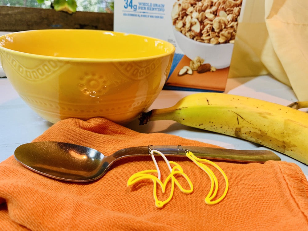 In the background there is a box of cereal, a bright yellow bowl, and a ripe banana. In the front there is a orange cloth napkin with a spoon resting on it. On the handle of the spoon are two R+D earrings. They are mismatched. One is a yellow banana that has been peeled down to reveal the white fruit. The other is a unpeeled yellow banana. 