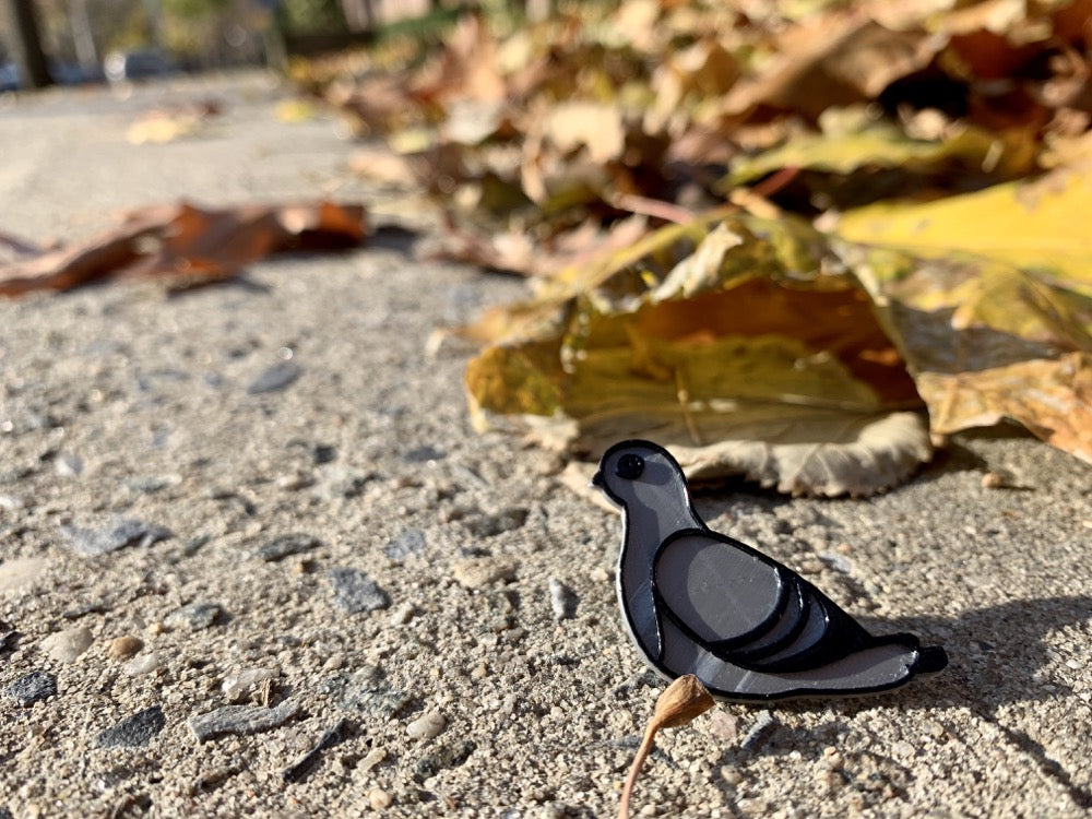 Resting on the sidewalk outside a park is a close up of a 3D printed pin. It is silver and black, shaped like a pigeon and 3D printed with plant based materials. 