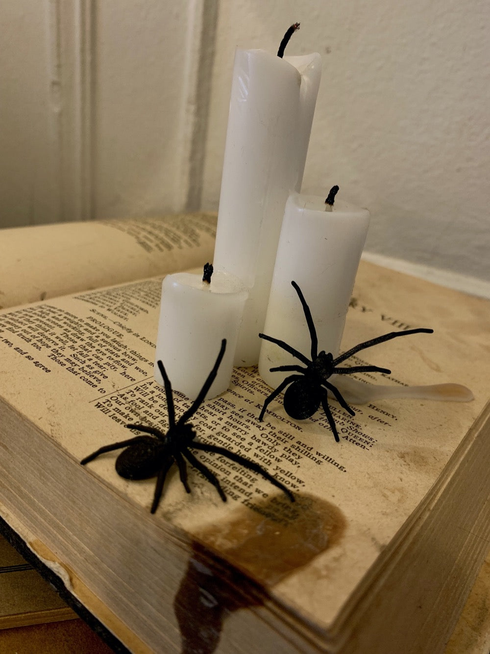 Shown are two black spider earrings that are realistic in their shape. They have long front legs that look like they are searching for their next bit of prey. They are pictured crawling up an old book with candles on top to create a spooky Halloween scene.