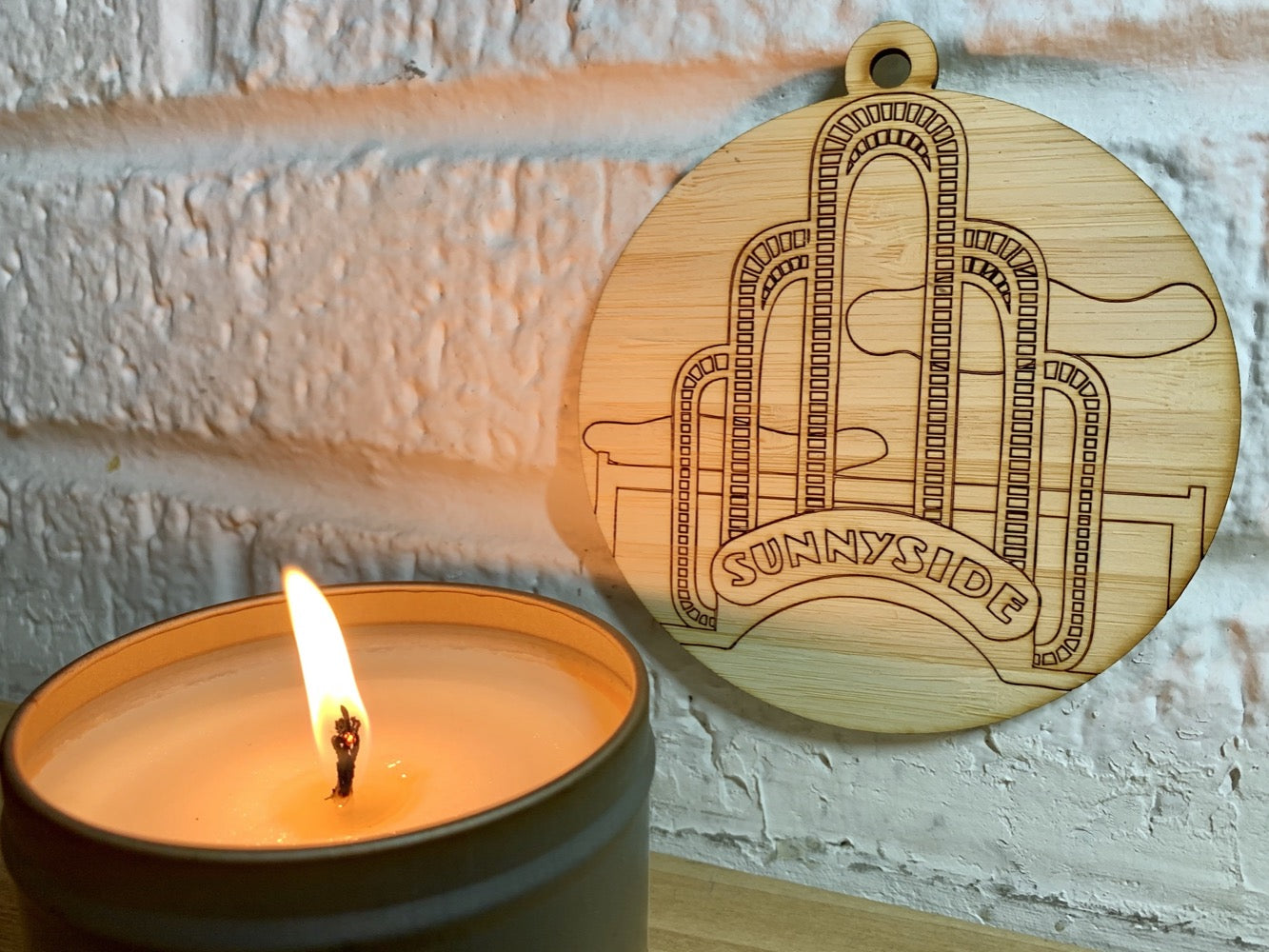 Shown on a white brick wall and next to a candle is a laser cut wall hanging. It is cut from bamboo, a fast growing and sustainable grass. Etched in it a picture of the iconic Sunnyside Arch in the center of the Queens neighborhood. Each piece can be painted like a paint by number or left plain. It can hang from your Christmas Tree or on you wall all year round.