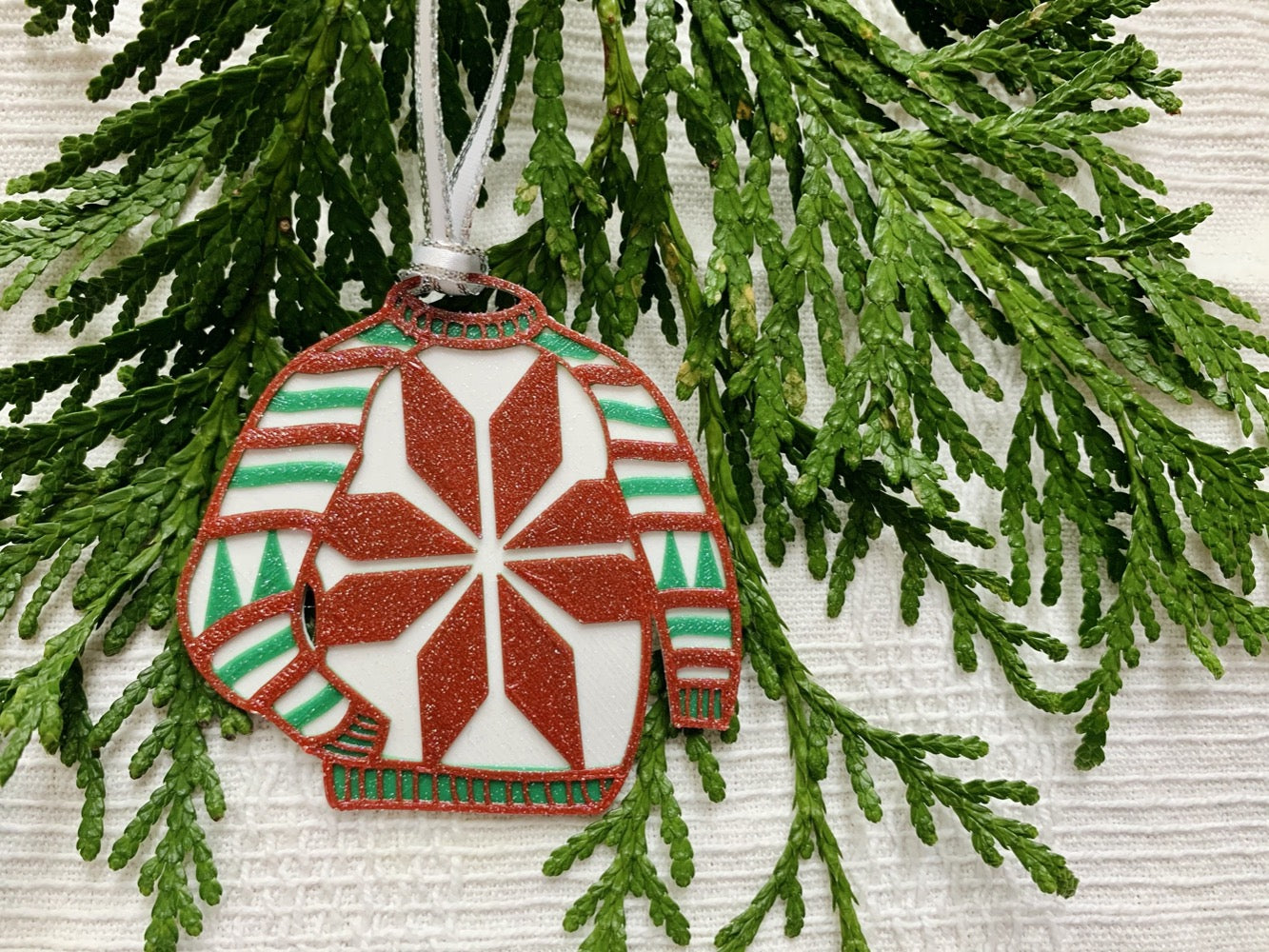 Shown on a white fabric background and with sprigs of evergreen is a R+D 3D printed ornament. It is in the shape of a chunky holiday sweater. It is white with red and green stripes, trees on the sleeves, and a snowflake in the center. The entire ugly sweater ornament is covered in glitter so it will shimmer and shine in the light. 