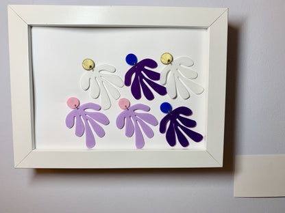 Within a white frame with a small card next to it (suggesting the set up of a museum) are three pairs of earrings. The R+D earrings have a circlar piece at the top linked to an abstract shape based on Matisse's cut out work. The earrings are in gold and white, blue and purple, and light pink and light purple. 
