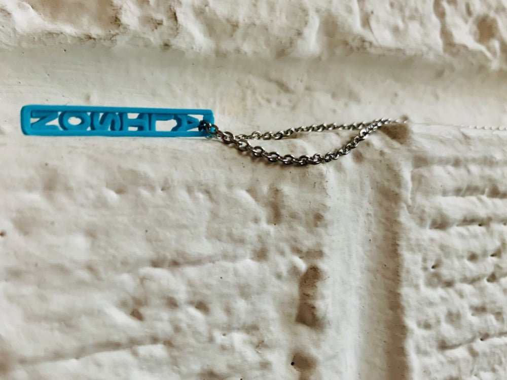 Laying on a white brick ledge is a necklace with a 3D printed pendant. The pendant is teal and have the name ALISON printed in it. These pendants can be personalized to any name, word, or date. 