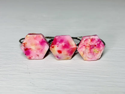 On a white background are three rings side by side. They are each  have a hexagonal shaped charm on the front of the band. The first is a smooth hexagon, the second a faceted hexagon, and the third a bumpy geode. They are cast from recycled 3D prints in red, yellow, orange, hot pink, and white. 