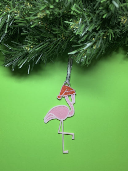 On a bright green background with a wreath above it is a R+D 3D printed ornament. It is a light pink flamingo that has one leg up and is wearing a red santa hat.