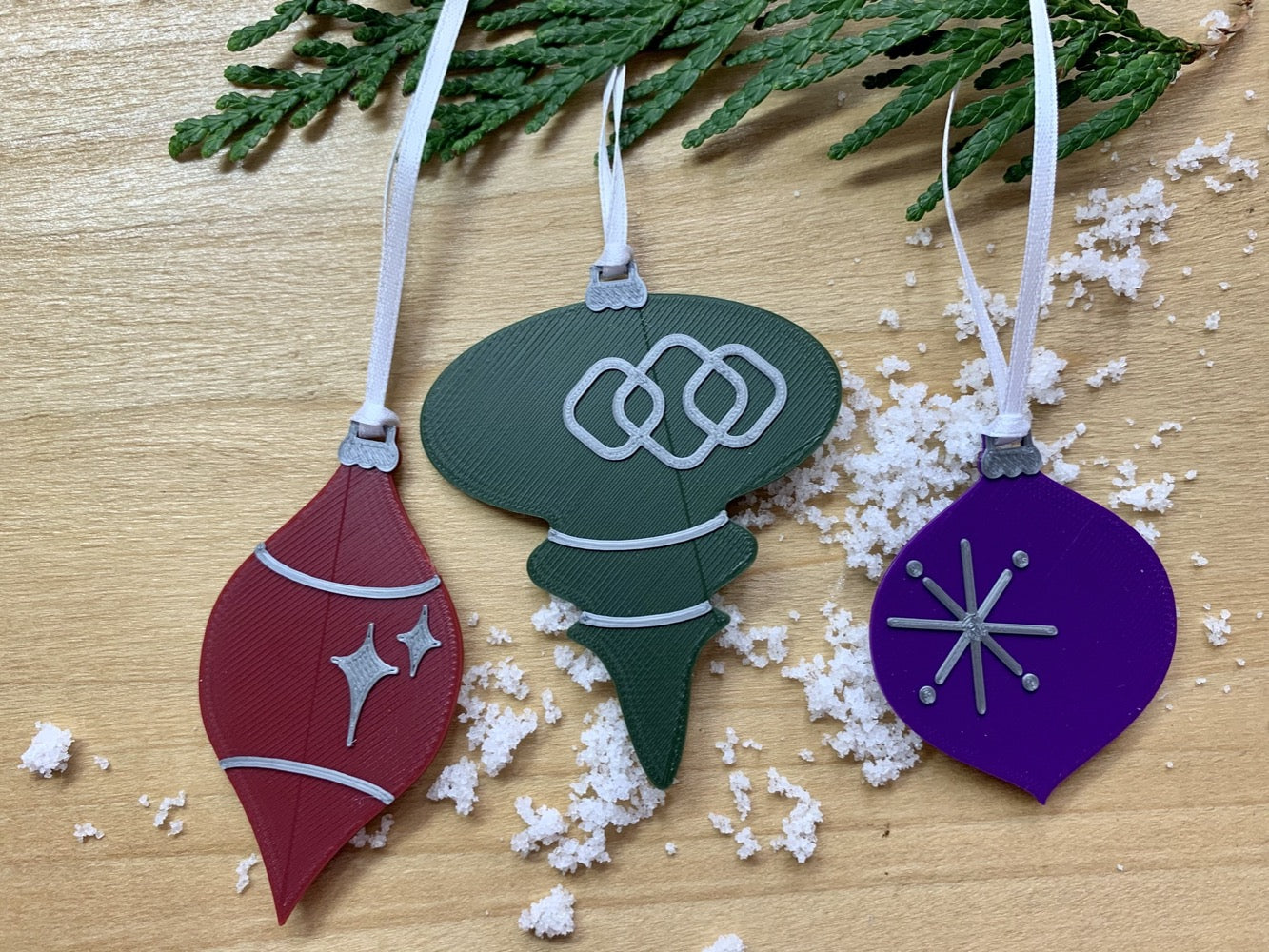 On a wood background with white snow and hanging from an every green branch are a set of 3 R+D 3D printed ornaments. They are all designed to look like vintage baubles that would have commonly been found on Christmas trees in the past. They each have silver accents of stars and shapes. One is merlot red, one is dark green, and one is purple.